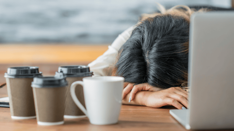 an exhausted worker slumps face down on her desk in front of her laptop surrounded by coffee cups
